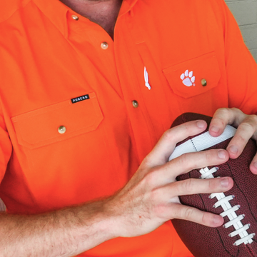 Man wearing bright orange shirt holding a football, featuring The All In logo on chest pocket