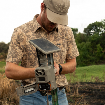 Man wearing "The Armadillo" patterned shirt and hat adjusts a solar-powered outdoor device in a natural setting.