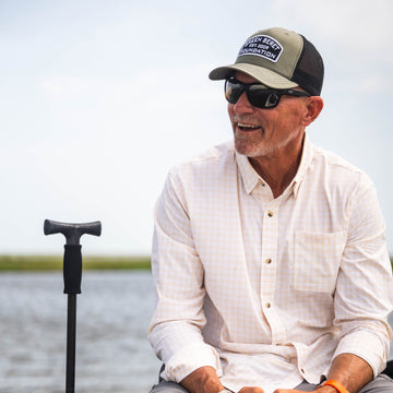 Man wearing sunglasses and a hat smiling outdoors by water, dressed in The Baja shirt.