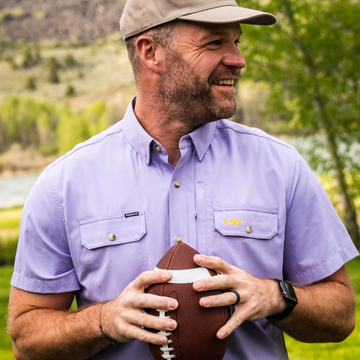 Man outdoors holding football wearing a lavender button-up shirt called The Baton Rouge with a cap and smartwatch