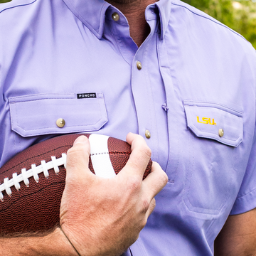 Close-up of a person wearing The Baton Rouge shirt holding a football with LSU embroidery on the pocket.