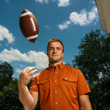 Man in an orange button down shirt reaching toward a football under a blue sky outdoors