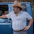 The Big Rig wearing a striped shirt and cowboy hat beside a blue pickup truck