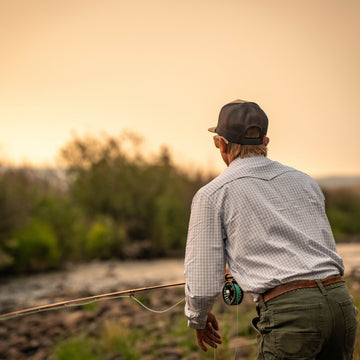 The Bighorn shirt worn by an angler holding a fly rod beside a river at sunset