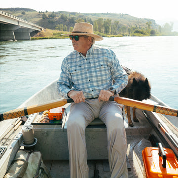 Man wearing The Blue Bison shirt rowing a boat on a river with a dog and outdoor gear nearby