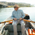 Man wearing The Blue Bison shirt rowing a boat on a river with a dog and outdoor gear nearby