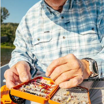 Close-up of a person organizing colorful fishing flies while wearing The Blue Bison shirt outdoors near water.