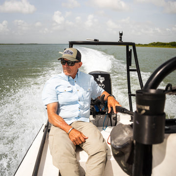 Man wearing sunglasses and a cap sitting on a boat wearing a light blue Blue Steel shirt on a sunny day outdoors