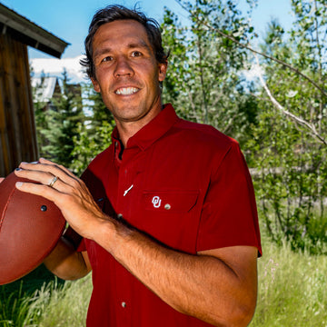 Man in a red button-up shirt holding a football outdoors near trees