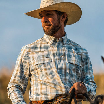 Man wearing The Brush Country shirt and cowboy hat outdoors, holding reins against a clear sky background