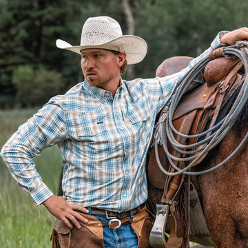 Man wearing The Brush Country shirt and cowboy hat leaning on a saddled horse in an outdoor setting