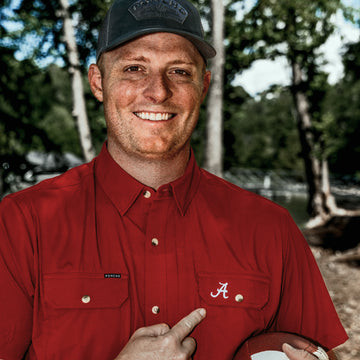 Smiling man in a red button up shirt and dark cap pointing to an embroidered letter A outdoors