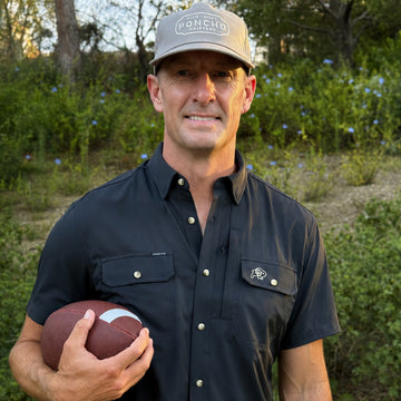 Man wearing a black button-up shirt and cap, holding a football outdoors