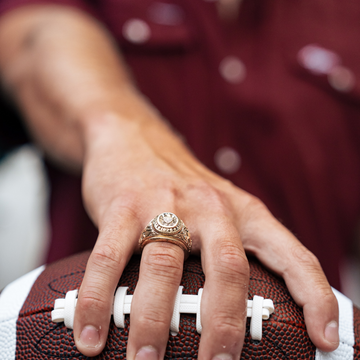 Close-up of a hand wearing The College Station ring resting on a football with a maroon shirt in the background