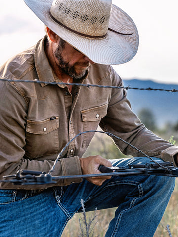 Man wearing The Cotulla shirt and cowboy hat working with wire fencing in an outdoor rustic setting