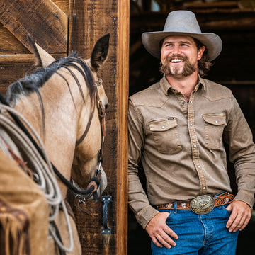 Man wearing a wide-brim hat and The Cotulla shirt smiling next to a horse at a wooden barn door