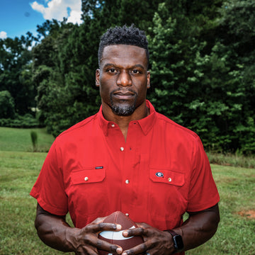 Man wearing a red button-up shirt holding a football outdoors in a grassy field