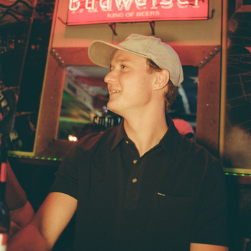 Young man wearing a black shirt and beige cap smiling indoors under a Budweiser sign, featuring The Front Porch shirt.