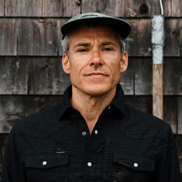 The Grey Beard man wearing a dark plaid shirt and cap against a rustic wooden wall