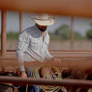 Man wearing The Hammer Down shirt and cowboy hat, preparing gear behind metal fence outdoors