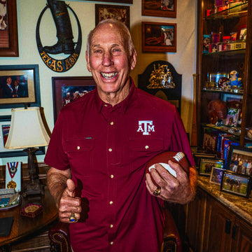 Smiling older man in a maroon A&M shirt holding an American football in a memorabilia-filled room