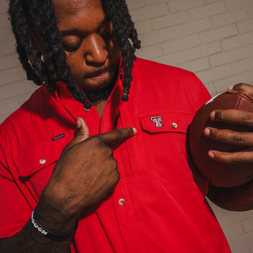 Man in a red button-up shirt pointing to a Texas Tech logo while holding an American football