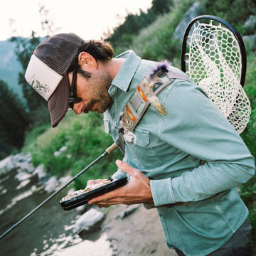 Man wearing The Mentone shirt while fishing near a river, holding fishing gear and wearing a cap and sunglasses