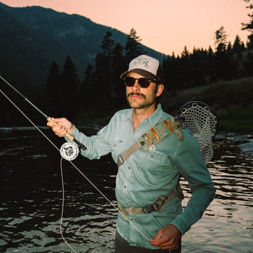 Man wearing The Mentone shirt and sunglasses fly fishing at dusk by a river with mountains and trees in the background