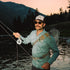 Man wearing The Mentone shirt and sunglasses fly fishing at dusk by a river with mountains and trees in the background