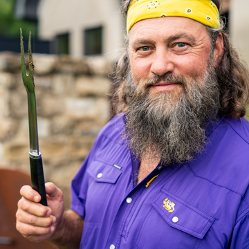 Man wearing a yellow bandana and "The Mike" purple shirt holding a grilling fork outdoors near a stone wall.