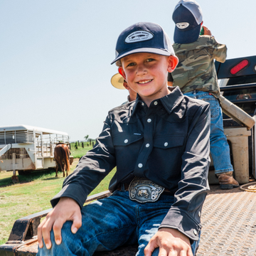 Young boy wearing a dark button-up shirt and hat sitting outdoors by a truck, showcasing The Newt shirt.