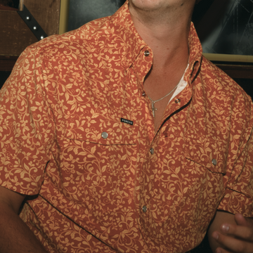 Close-up of a person wearing The Occupy Left Field shirt with an intricate yellow floral pattern on rust background outdoors style.