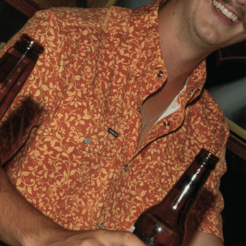 Close-up of a smiling man wearing The Occupy Left Field shirt with an intricate orange floral pattern, holding a dark bottle.