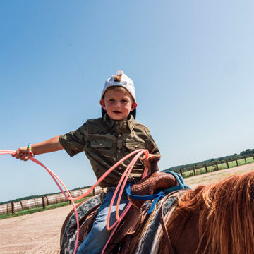 Young boy wearing The Palomita shirt while riding a horse outdoors under a clear blue sky