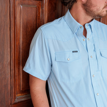 The Pedernales light blue button up shirt worn by a man beside a wooden door