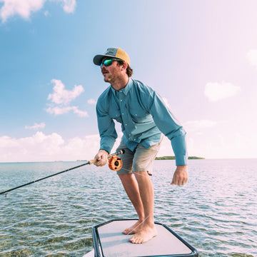 Man in blue shirt and cap fishing barefoot on boat in clear water under bright sky