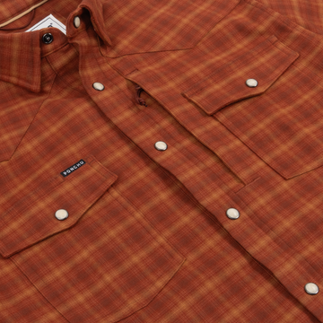 Close-up of a rust-colored plaid shirt with snap buttons and chest pockets featuring a small "The Pitstop" label tag.