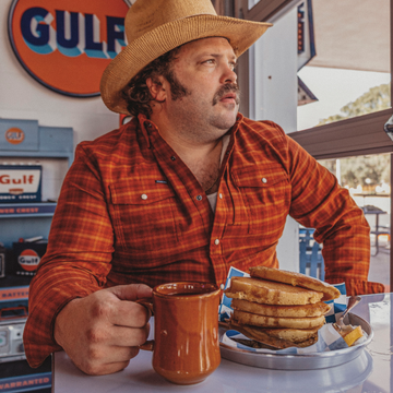 The Pitstop man in a red plaid shirt holding coffee beside a stacked toasted sandwich at a diner