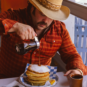 The Pitstop man in a red plaid shirt pouring syrup over a stack of pancakes