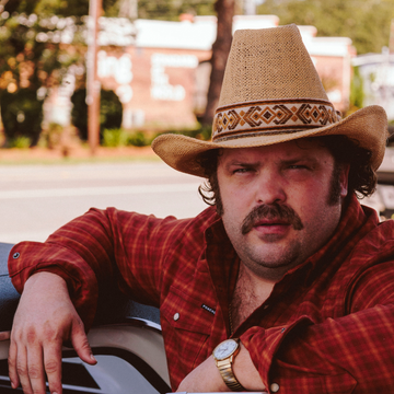 The Pitstop featuring a man in a cowboy hat and red plaid shirt leaning against a vehicle