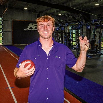 Smiling man in a purple shirt holding a red ball inside an indoor training facility