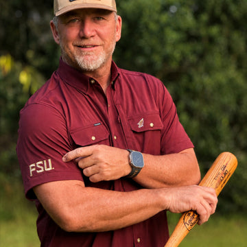 Man wearing a burgundy short-sleeve shirt named The Seminole with FSU embroidery on sleeve outdoors holding a bat