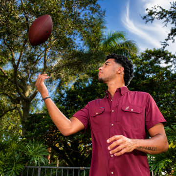 Man wearing The Seminole shirt tossing a football outdoors against a backdrop of trees and blue sky