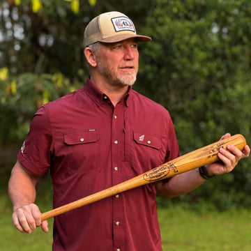 Man outdoors wearing The Seminole shirt in deep red holding a wooden baseball bat in a natural setting