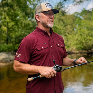 Man wearing The Seminole shirt while fishing near a river surrounded by green trees.