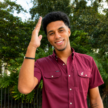 Smiling man in a burgundy short-sleeve button shirt raising his hand outdoors