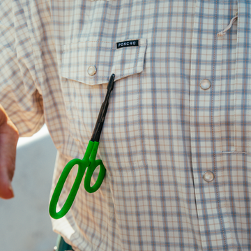 Close-up of a plaid shirt pocket labeled The Soda Butte with green-handled scissors clipped to it outdoors