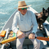 Man wearing a straw hat and The Soda Butte shirt rowing a boat with a black dog nearby on the water