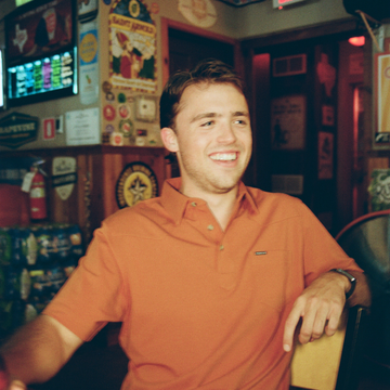 Young man smiling and sitting in a cozy bar wearing an orange polo shirt with relaxed ambiance in the background