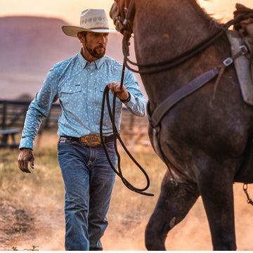 Man wearing The St. Helens shirt and a cowboy hat while leading a horse in an outdoor setting at sunset
